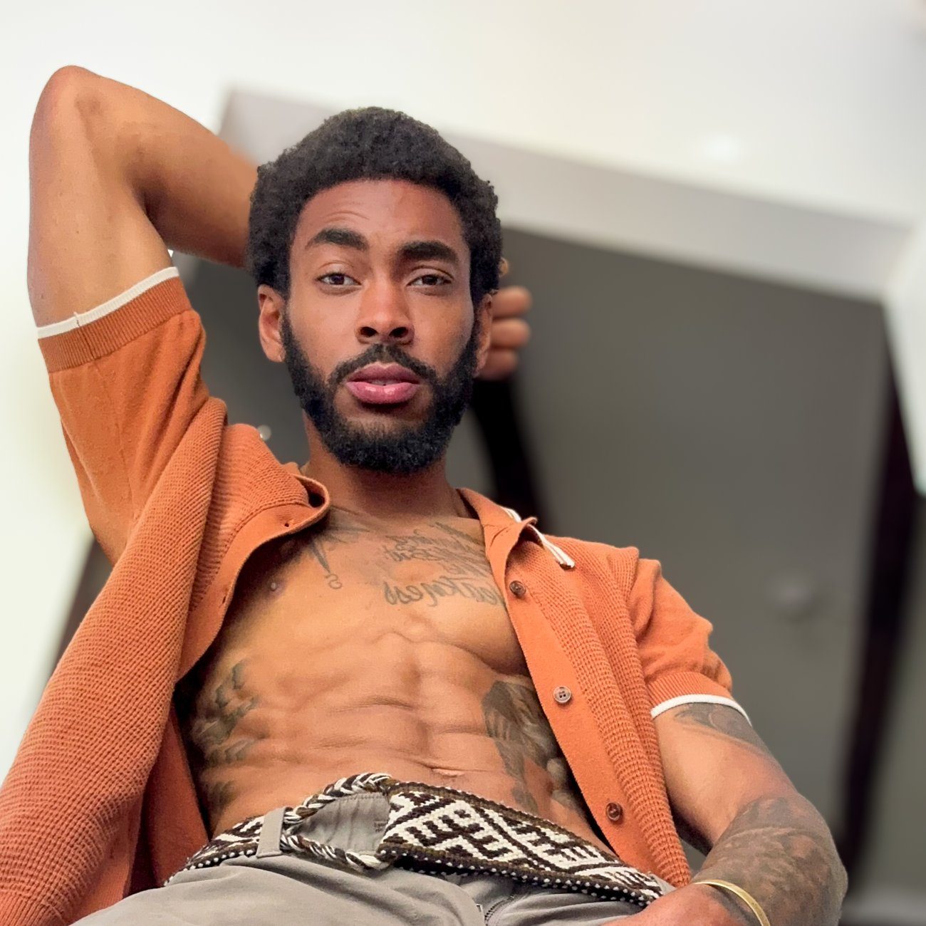 Zen leaning back in an open rust coloured shirt, one arm behind his head, holding the camera with a steady unhurried look.